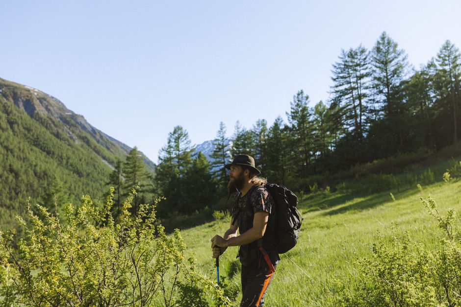 A man with a backpack hikes in a lush green mountain landscape under a clear blue sky.