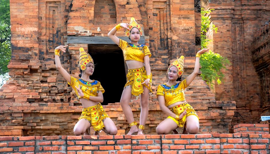 Three dancers in traditional costumes performing at a historic temple with intricate brickwork.