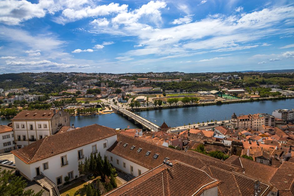 Aerial view of Coimbra, Portugal, showcasing the Mondego River and historic architecture under a blue sky.