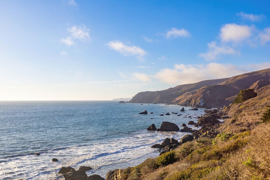Beautiful view of a rocky coastline with waves and hills under a clear blue sky.