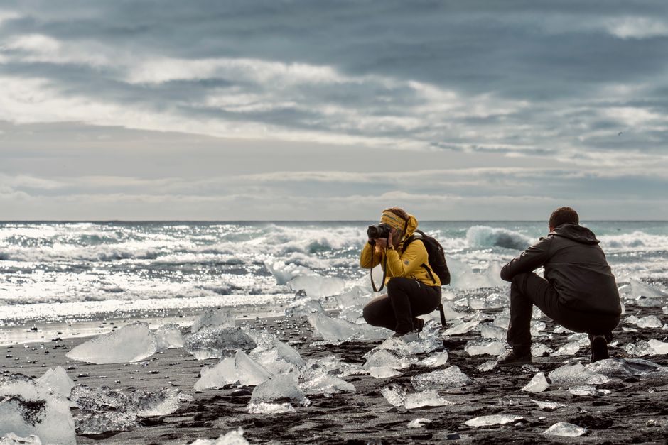 Two photographers capturing stunning ice formations on a black sand beach by the ocean.