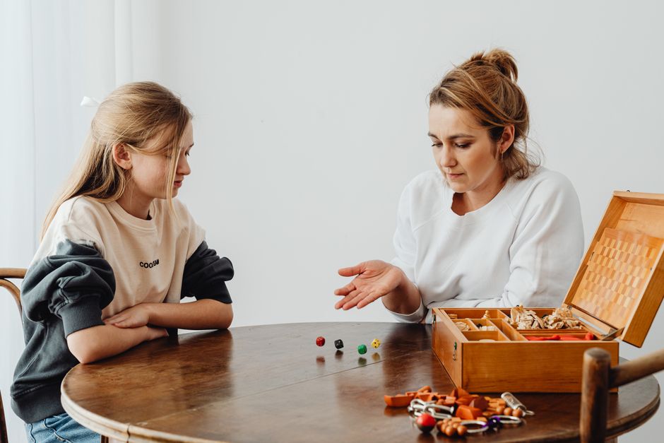 A mother and daughter enjoy a board game indoors, surrounded by dice and game pieces.