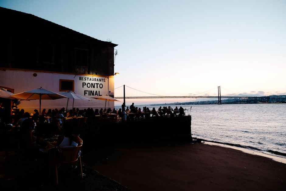 Scenic view of people dining outdoors at Restaurante Ponto Final with Lisbon's sunset and bridge in the background.