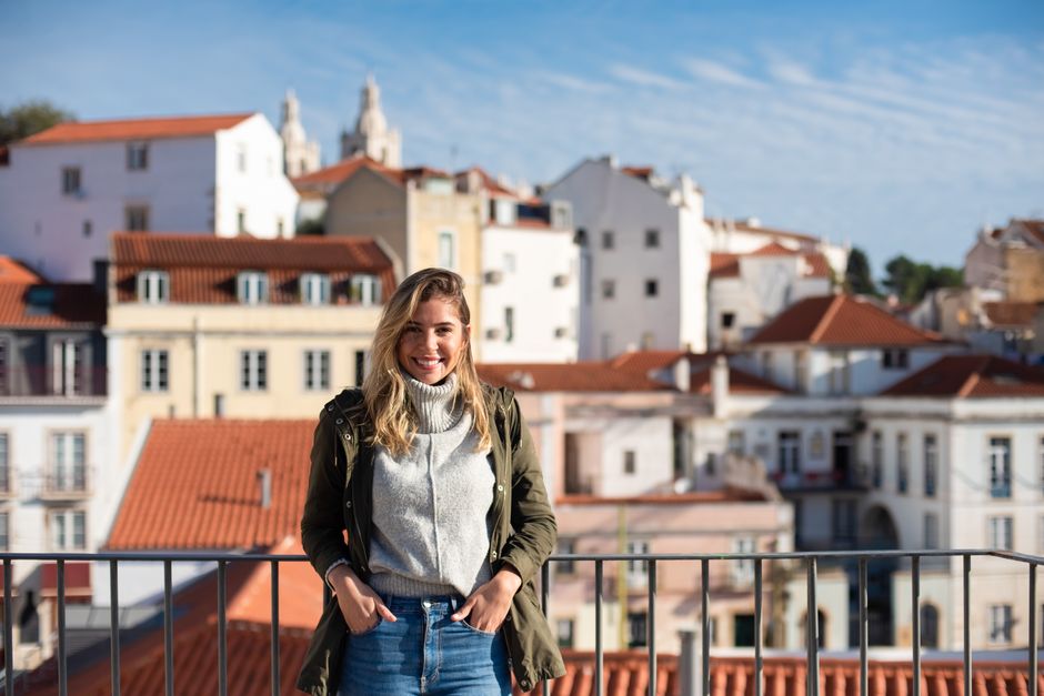 Young woman in turtleneck smiling on a balcony overlooking Lisbon's historic architecture.