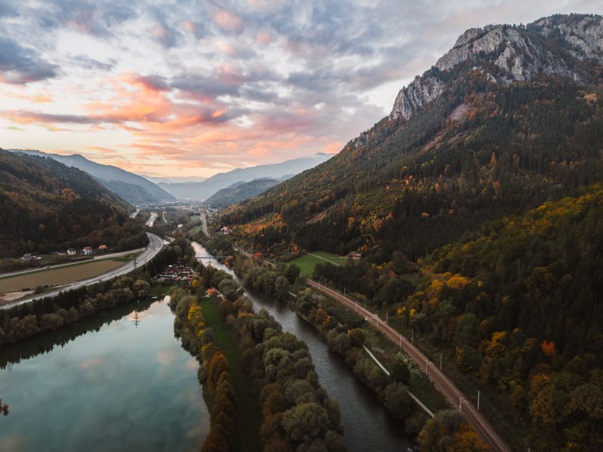 A breathtaking aerial view of the Austrian mountains at sunset with a vibrant sky and mirrored river below.