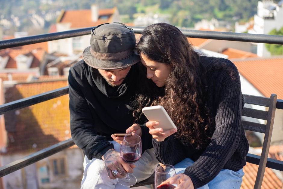 A couple enjoying a sunny day with smartphones and wine glasses on a rooftop in Portugal.