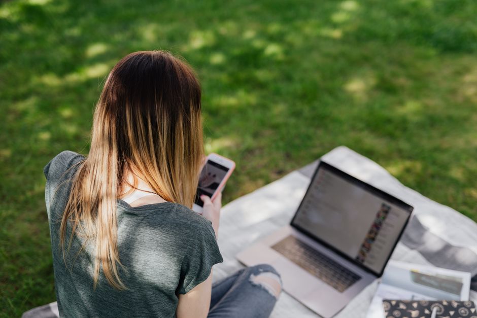From above back view of skinny long haired teenage girl in casual clothes browsing social media on smartphone while spending time in sunny park during summer vacation