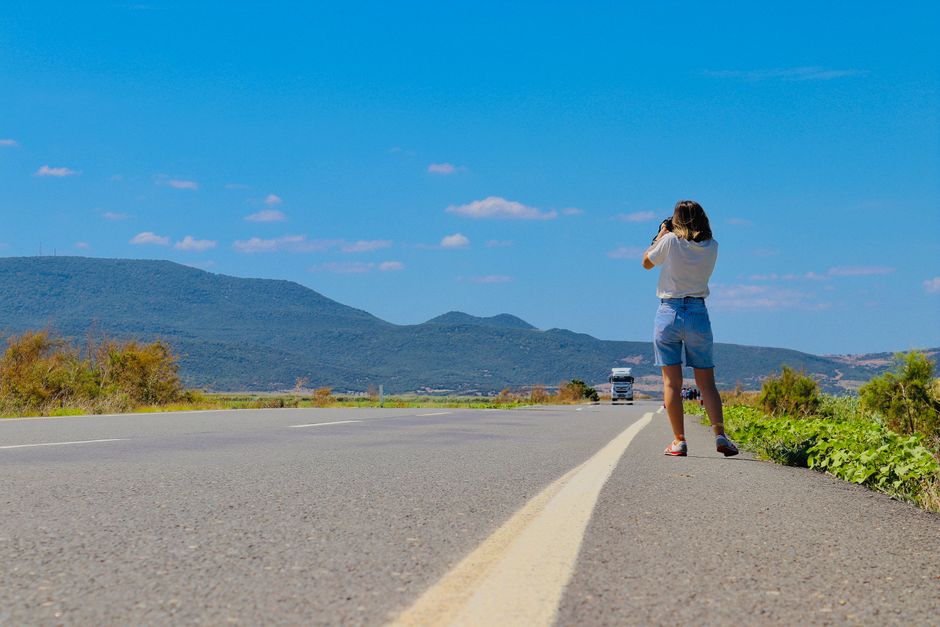 A woman in denim shorts captures the scenic beauty of mountains along an open road under a clear blue sky.