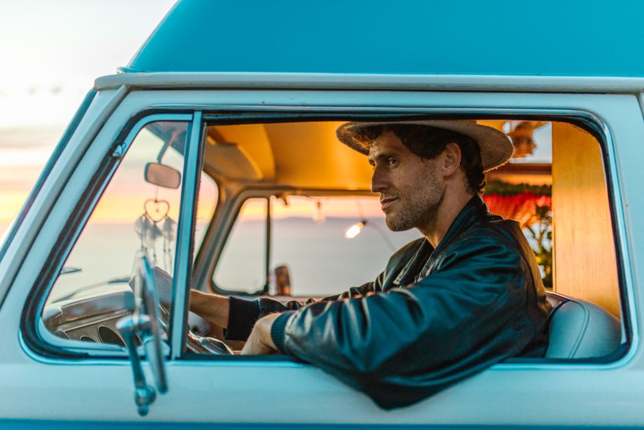 A man in a hat drives a classic camper van with a sunset view.
