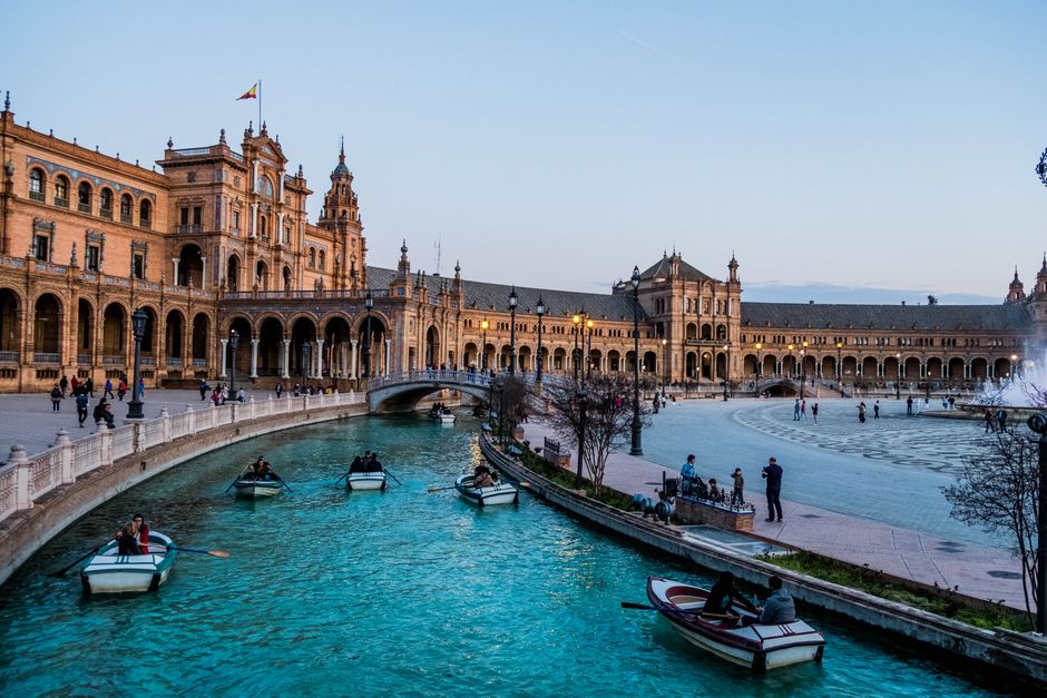 Boats on the canal at Plaza de España, Seville, with historic architecture and vibrant sky.