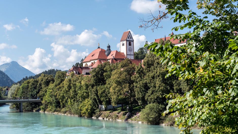 Picturesque Saint Mang Monastery in Füssen, Bavaria, framed by lush trees and river.