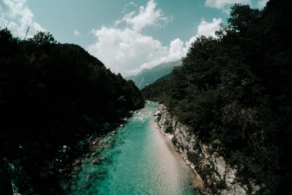 From above of green rippled river between high mountains with trees under sky with clouds
