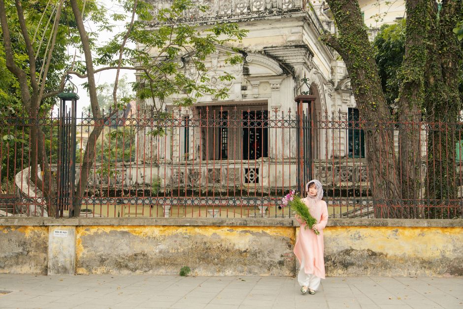 A young woman in traditional attire holding flowers in front of a historic building in Hanoi, Vietnam.
