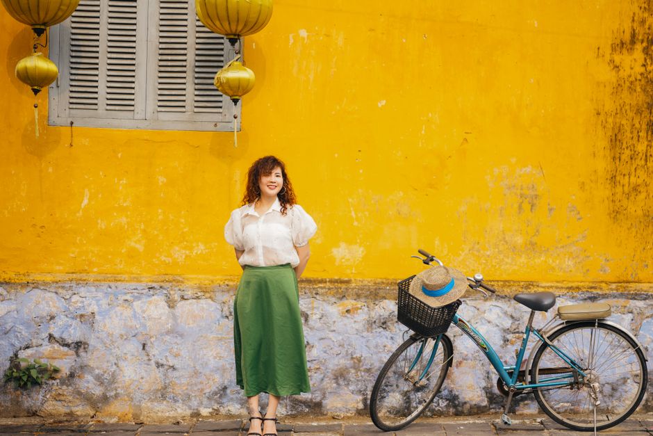 Asian woman in green skirt smiling beside bicycle and yellow wall.