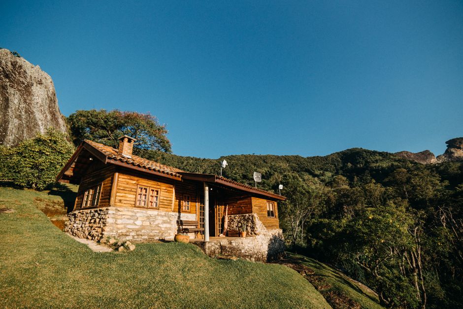 Aged hut located on grassy hill near rock and dense forest with green trees in suburb area in countryside on summer day