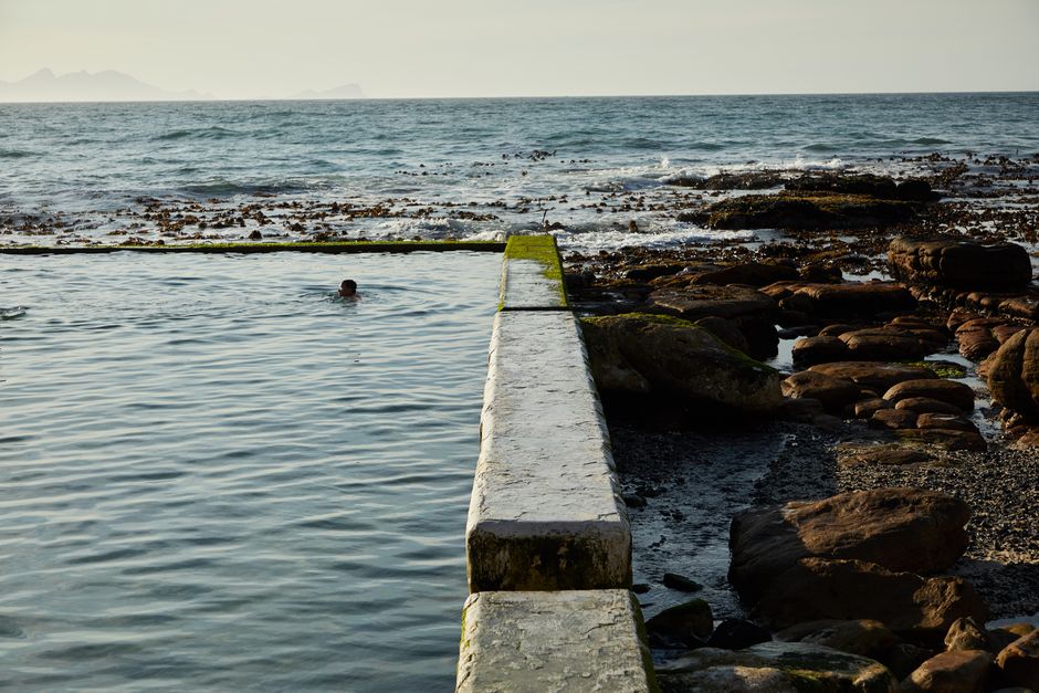 A person swims in a tidal pool by the rocky sea, with crashing waves in the background. Daytime setting.