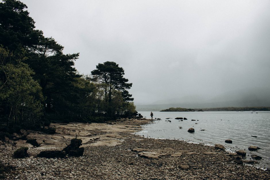 Moody lakeside view with fog, rocky shoreline, and distant figure adds serene atmosphere.