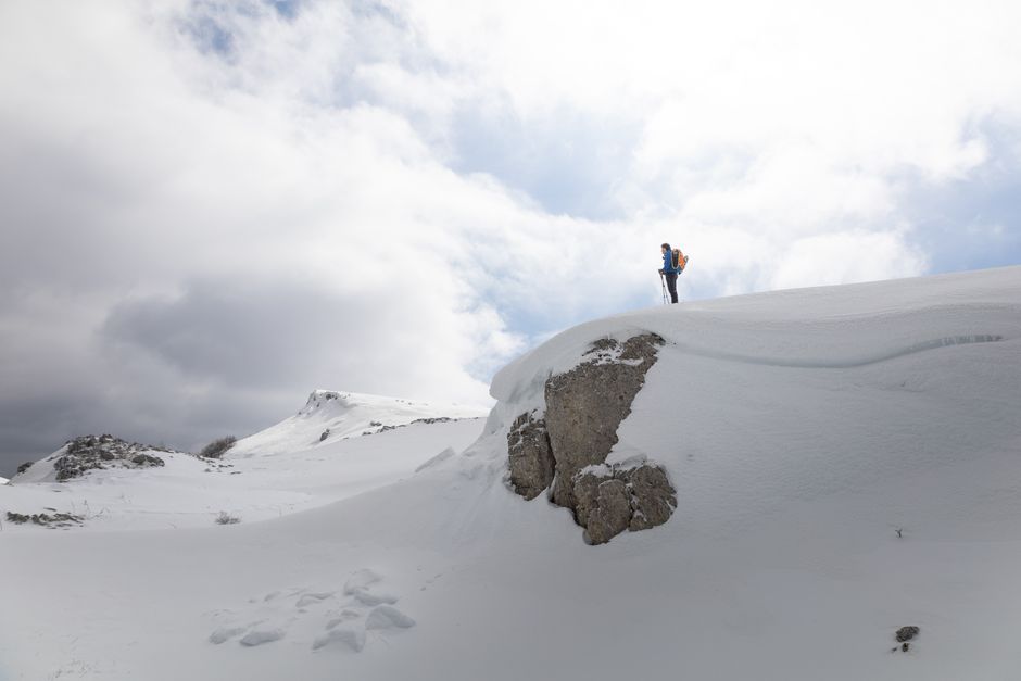 A solitary hiker standing on a snow-covered mountain peak in Abruzzo, Italy under a dramatic sky.